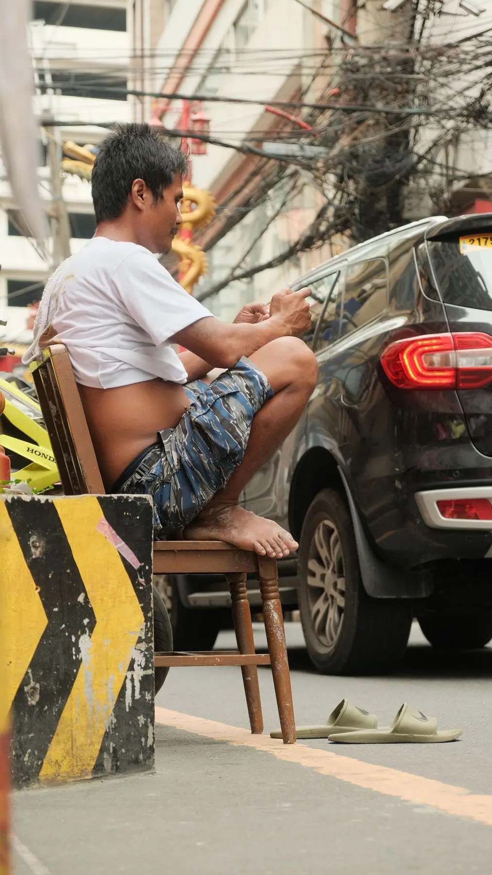 Lalaking nakaupo sa bangkito sa kalsada sa Maynila, nakatingin sa telepono, may mga kawad sa itaas ng kalye.