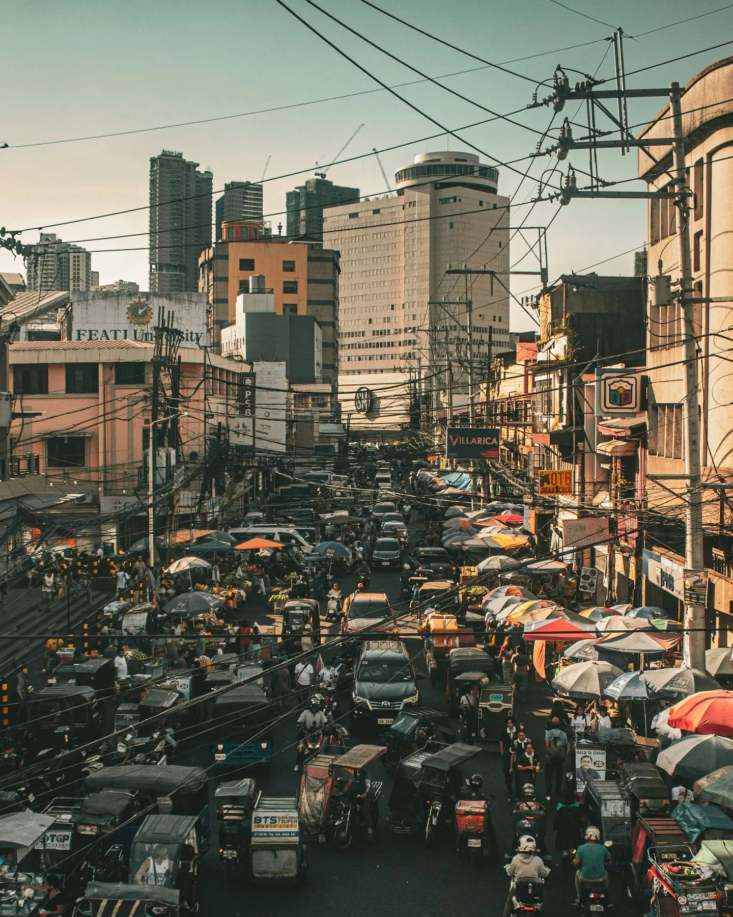 Siksik na kalye sa Maynila na may traysikel, payong, at matataas na gusali sa malayo.