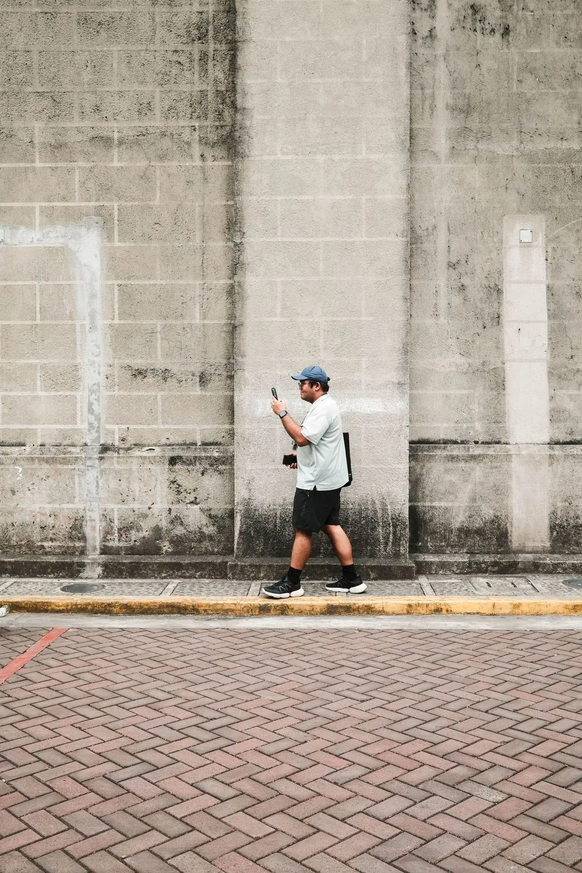 Lalaking naglalakad sa Maynila habang tinitignan ang telepono sa tabi ng konkretong pader.